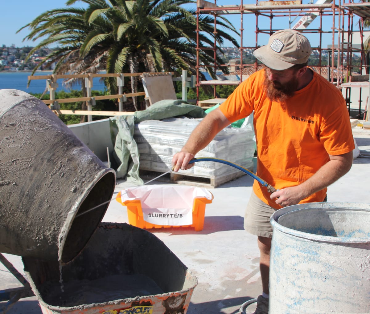 Man in hat on a sunny construction site with palm trees in the background rinsing a concrete mixer into a slurrytub.